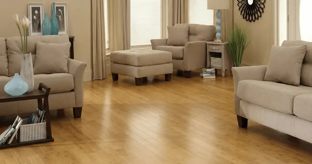 A living room with professionally installed bamboo flooring, beige sofas, a matching ottoman, a side table with a blue lamp, vases, framed pictures, and large windows with blinds and curtains in Miami, FL